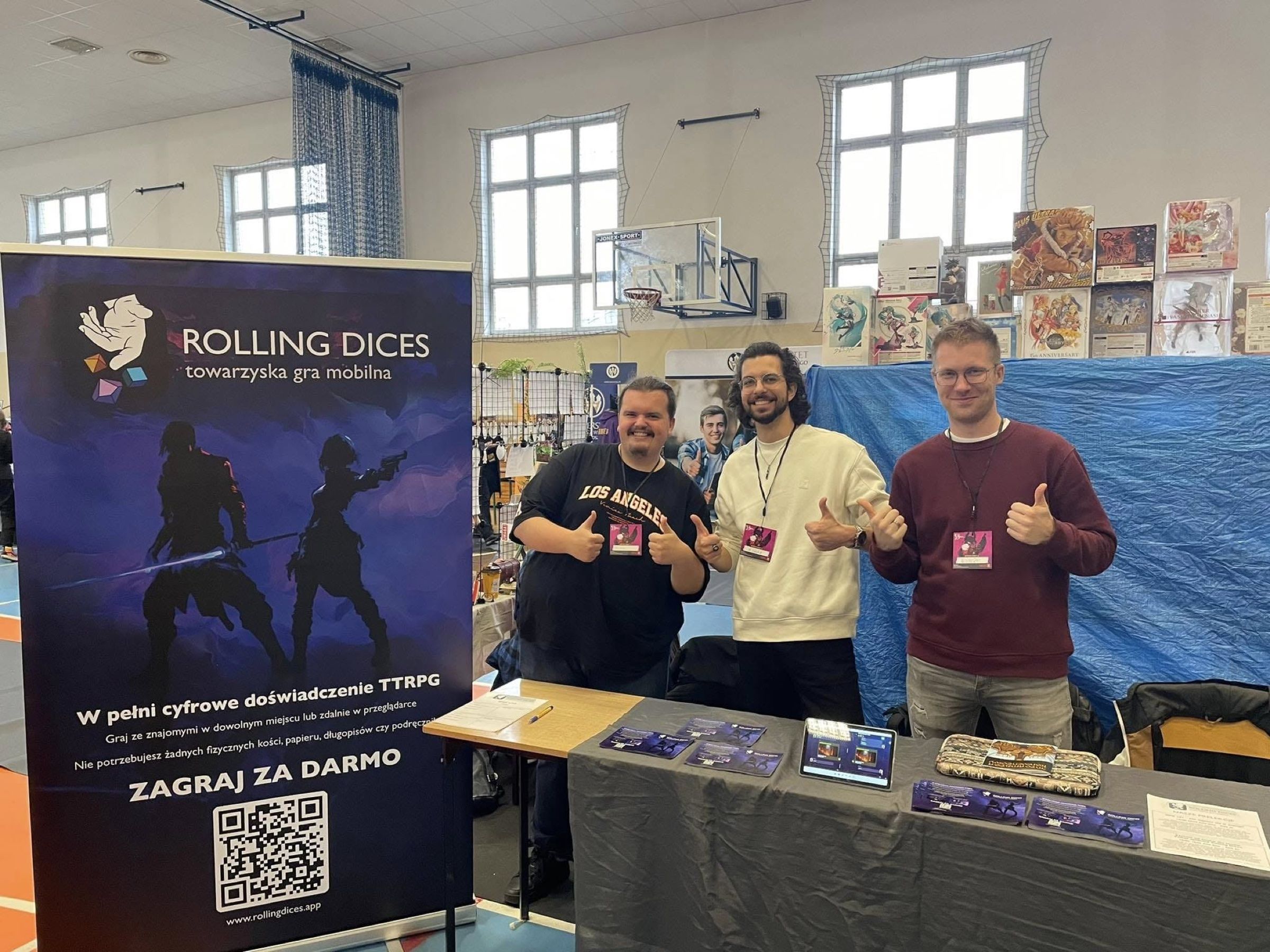 Three team members behind a convention stand with a Rolling Dices roll-up banner, flyers and a tablet running the app.